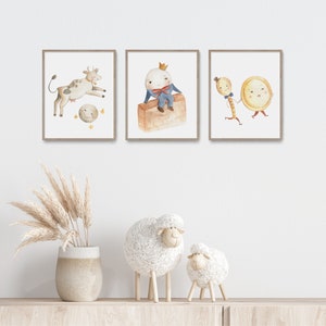 May include: Three framed watercolor prints featuring nursery rhyme characters: a cow jumping over the moon, Humpty Dumpty, and a spoon and plate. Below, two decorative sheep figurines and a vase with dried plants sit on a wooden shelf.