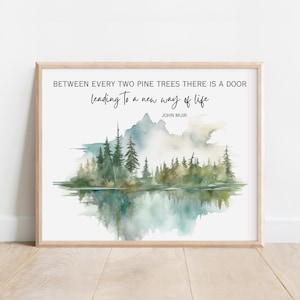 May include: A framed art print featuring a watercolor landscape with pine trees, a lake, and mountains. The text reads, "BETWEEN EVERY TWO PINE TREES THERE IS A DOOR leading to a new way of life" by John Muir. The frame is light brown.