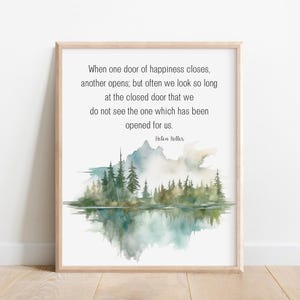 May include: A framed wall art print featuring a quote by Helen Keller in black text above a watercolor illustration of a forest landscape with green pine trees, blue mountains, and a reflective water surface in soft muted tones.