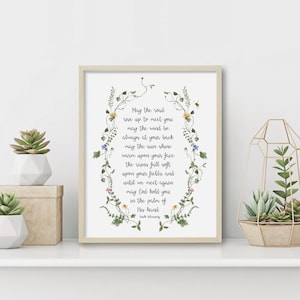 May include: A framed print with a white background and a floral border. The print features a poem about a road, wind, sun, rain, and fields. The poem reads: "May the road rise up to meet you, may the wind be always at your back, may the sun shine warm upon your face, the rains fall soft upon your fields, and until we meet again may God hold you in the palm of His hand." The poem is attributed to Irish poet Frank O'Hara.