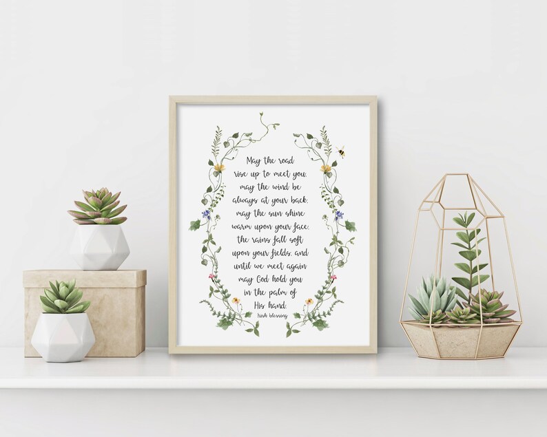 May include: A framed print with a white background and a floral border. The print features a poem about a road, wind, sun, rain, and fields. The poem reads: "May the road rise up to meet you, may the wind be always at your back, may the sun shine warm upon your face, the rains fall soft upon your fields, and until we meet again may God hold you in the palm of His hand." The poem is attributed to Irish poet Frank O'Hara.