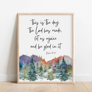May include: A framed wall art print featuring a watercolor landscape of evergreen trees against purple and orange mountains. The text reads: This is The day The Lord has made, let us rejoice and be glad in it. Psalm 118:24 in black script.