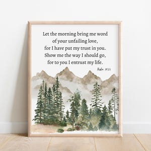 Op de afbeelding: Een ingelijste kunstdruk met een aquarel landschap van bergen en groenblijvende bomen. Het kunstwerk bevat de tekst "Let the morning bring me word of your unfailing love..." en "Psalm 143:8". De lijst is lichtbruin.