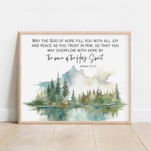 May include: A framed print featuring a watercolor landscape with a lake, trees, and mountains. The artwork includes the text "MAY THE GOD OF HOPE FILL YOU WITH ALL JOY AND PEACE..." and "The power of the Holy Spirit" from Romans 15:13.