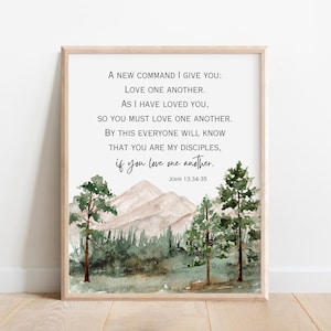 Può includere: Una stampa incorniciata con un paesaggio ad acquerello con montagne e alberi. La stampa mostra il testo "A new command I give you: Love one another..." da Giovanni 13:34-35. La cornice è di colore legno chiaro.