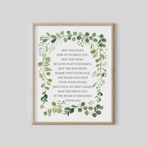 May include: Framed print featuring an Irish blessing surrounded by a watercolor wreath of green leaves. The text reads "May the Road Rise Up to Meet You..." in a classic serif font. A light wood frame surrounds the print.