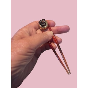 Peut inclure: Un accessoire de cheveux vintage, avec un dessus décoratif doré avec un motif noir et or. Le bâton à cheveux a un design mince et pointu, de couleur brun rougeâtre. Le fond est rose clair uni.