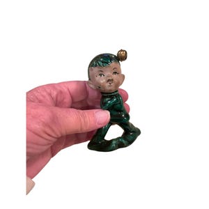 May include: A vintage ceramic elf figurine, depicted walking, with green attire and hair. The elf has a painted face and a small gold bell on its head. The figurine is held in a hand against a white backdrop.