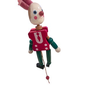 May include: A wooden pull-string toy in the shape of a rabbit. The rabbit has a white head with blue eyes, a red nose, and pink ears. It wears a red vest with a white tie and green arms and legs. The toy is painted with a cheerful expression.