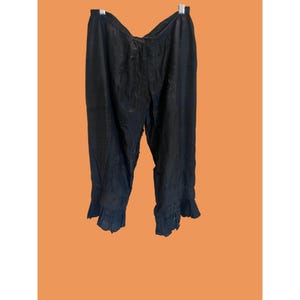 May include: Black, flowing trousers with ruffled hems hang against an orange background. The trousers are made of a lightweight fabric and appear vintage or bohemian. They are hanging from a metal hanger.