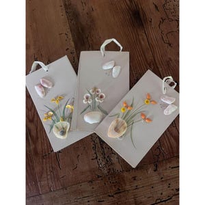 May include: Three seashell art pieces with floral designs. Each piece features a beige background with a painted vase, flowers, and seashells. White ribbon loops are attached for hanging. The seashells are in shades of pink, white, and orange.