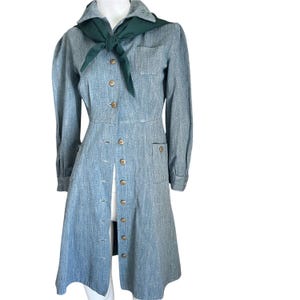 May include: A vintage light blue dress with long sleeves, a collared neckline, and a button-down front. The dress features a dark green scarf tied at the neck and two front pockets. The dress has a tailored fit.