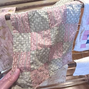 Puede incluir: Un edredón de retazos con una mezcla de cuadrados de tela estampada en rojo, gris y blanco. El edredón está colgado sobre un tendedero de madera, mostrando su construcción artesanal y estética vintage. También hay otros textiles colgando.