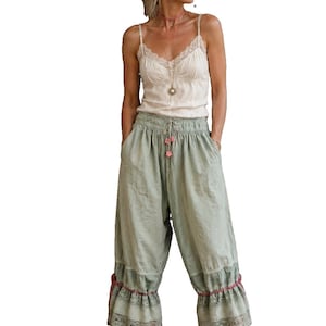 May include: A cream-colored camisole with lace trim at the neckline paired with sage green cropped trousers featuring a drawstring waist, pink floral accents, and a wide lace-trimmed ruffle at the hemline, worn with lace-up ballet flats.