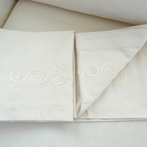 May include: Off-white bed sheets with a decorative wave pattern embroidered along the top edge. The sheets are folded neatly, showcasing the detailed design and the crisp, clean fabric. The bedding set appears to be new and unused.