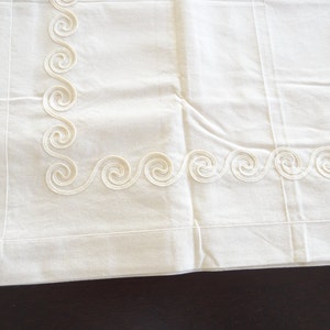 May include: Off-white pillowcase with a decorative border featuring a repeating swirl design. The pillowcase is made of a soft, possibly linen, fabric. The swirl pattern is a lighter shade of off-white.