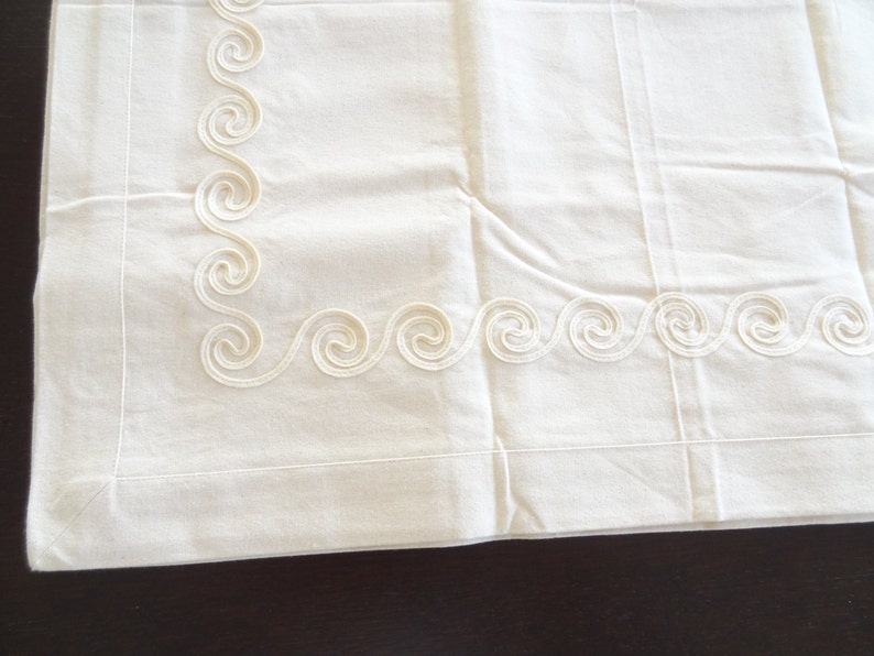May include: Off-white pillowcase with a decorative border featuring a repeating swirl design. The pillowcase is made of a soft, possibly linen, fabric. The swirl pattern is a lighter shade of off-white.