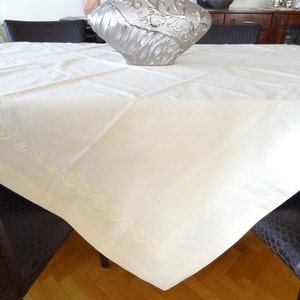 May include: Cream-colored square tablecloth with embroidered wave pattern along the edges. The tablecloth is draped over a table, with a decorative silver vase in the center. The table is surrounded by dark brown chairs.