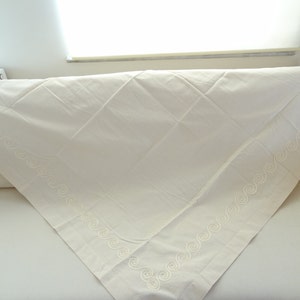May include: Cream-colored tablecloth with a decorative embroidered border featuring a repeating swirl pattern. The tablecloth is folded over a white surface, showcasing its texture and design. The fabric appears to be a soft, woven material.