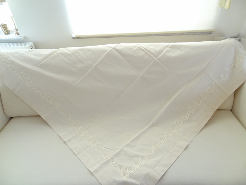 May include: Cream-colored tablecloth with a decorative embroidered border featuring a repeating swirl pattern. The tablecloth is folded over a white surface, showcasing its texture and design. The fabric appears to be a soft, woven material.