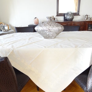 May include: A cream-colored tablecloth with embroidered details and a decorative silver vase on a dark wooden table. The tablecloth has a quilted pattern and a decorative border. The chairs around the table have a dark brown, textured upholstery.