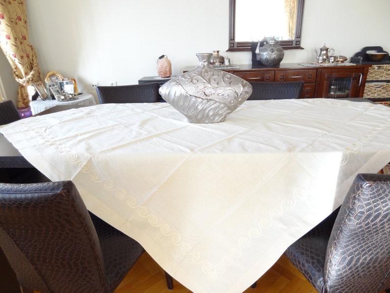 May include: A cream-colored tablecloth with embroidered details and a decorative silver vase on a dark wooden table. The tablecloth has a quilted pattern and a decorative border. The chairs around the table have a dark brown, textured upholstery.