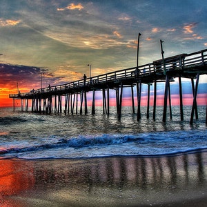 May include: A wooden pier extends out over a calm ocean at sunrise. The sky is a vibrant mix of pink, orange, and blue, with clouds reflecting the colors. The pier is supported by numerous wooden pilings, and there are a few people fishing on the pier.
