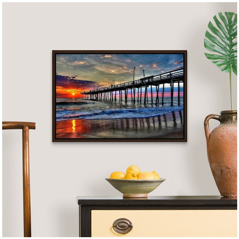May include: A colorful sunset over the ocean with a wooden pier extending into the water. The sky is a mix of orange, pink, and purple, and the water is a deep blue. The pier is silhouetted against the sky.