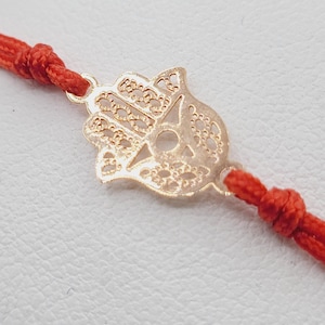 May include: A red string bracelet with a gold hamsa hand charm. The hamsa hand is a popular symbol of protection and good luck.