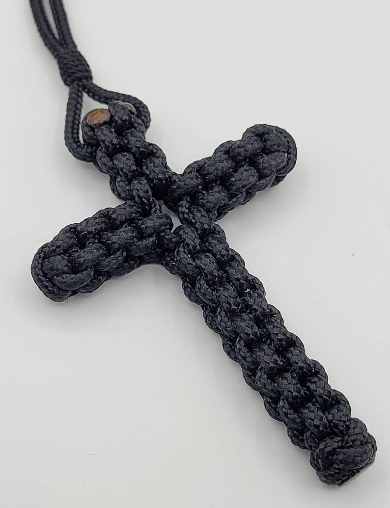 Puede incluir: Un colgante de cruz de cuerda anudada negra con una cuenta de madera marr&oacute;n en la parte superior.