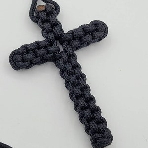 Puede incluir: Un colgante de cruz de cuerda negra anudada con una cuenta de madera marr&oacute;n en la parte superior.