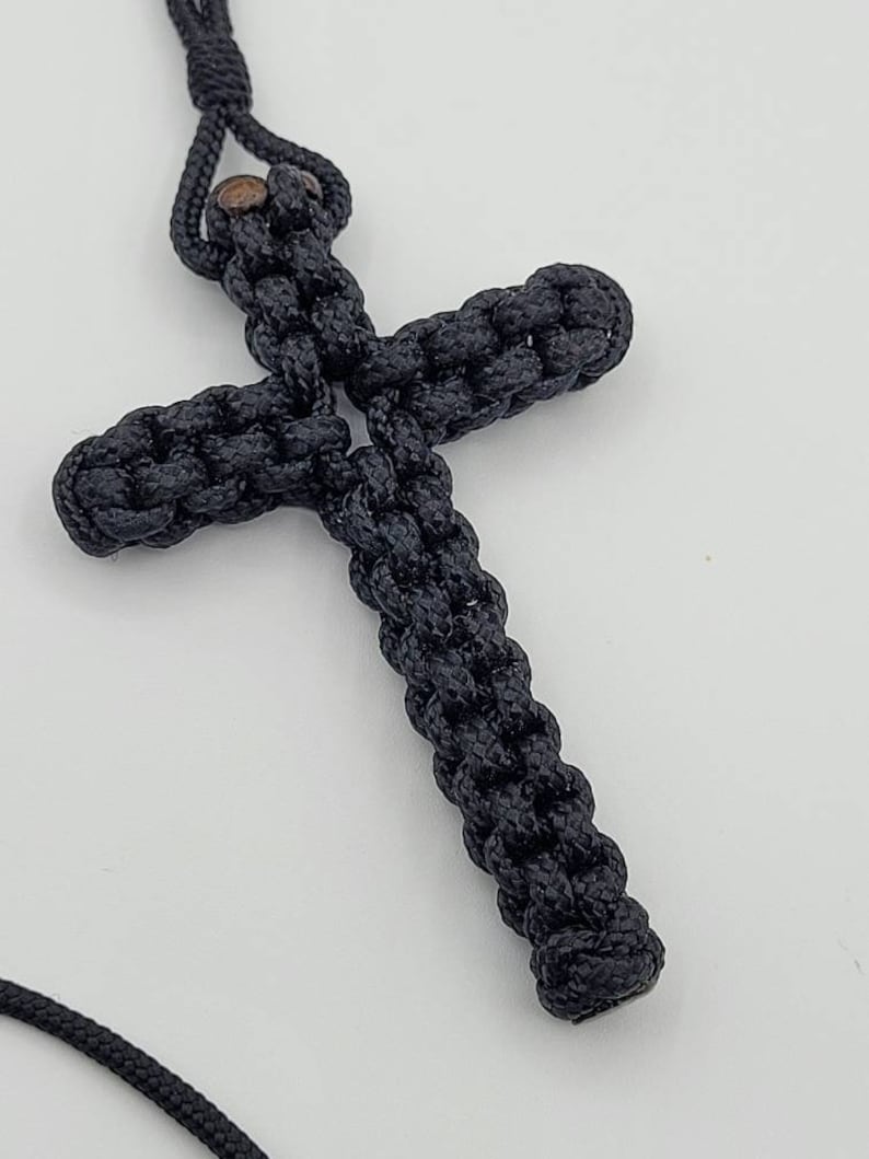 Puede incluir: Un colgante de cruz de cuerda negra anudada con una cuenta de madera marr&oacute;n en la parte superior.