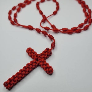 May include: A red knotted rosary with a red knotted cross. The rosary is made of a single strand of red cord with knots at regular intervals. The cross is a simple, symmetrical design.