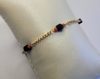 May include: A gold chain bracelet featuring alternating black faceted cube beads and small round orange beads. The bracelet is constructed with a delicate gold-tone metal chain, showcasing a repeating pattern of black and orange accents throughout.
