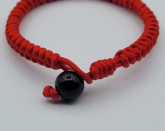 May include: Red string bracelet with a black bead. The bracelet is made of a braided cord and has a simple knot closure.