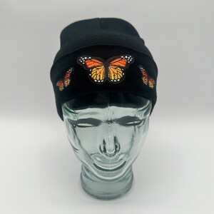 Peut inclure: Un bonnet noir avec trois motifs de papillons orange et noirs brodés.