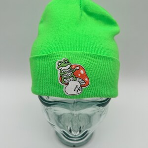 May include: A bright green beanie with a frog sitting on a mushroom embroidered patch. The frog is holding a guitar.