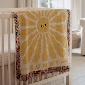 Könnte beinhalten: Eine gelb-cremefarbene gewebte Decke mit einem lächelnden Sonnendesign und dem Wort "SUNNY" am unteren Rand. Die Decke hat einen Fransenrand und ist über ein weißes Kinderbett drapiert. Das Kinderzimmer hat eine neutrale Farbpalette.