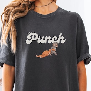 May include: Dark gray t-shirt with the text "team Punch" in a retro font. The shirt features an illustration of two cartoon characters. The shirt is a comfortable, casual style.