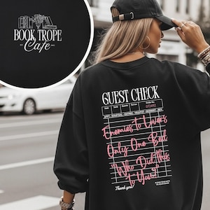 Könnte beinhalten: Schwarzes Sweatshirt mit dem Text "GUEST CHECK" und einer Menüliste. Darunter steht in rosa Schrift "Enemies to Lovers, Only One Bed, Who Did This to You?" Ein kreisförmiges Logo mit "BOOK TROPE Cafe" befindet sich oben links.