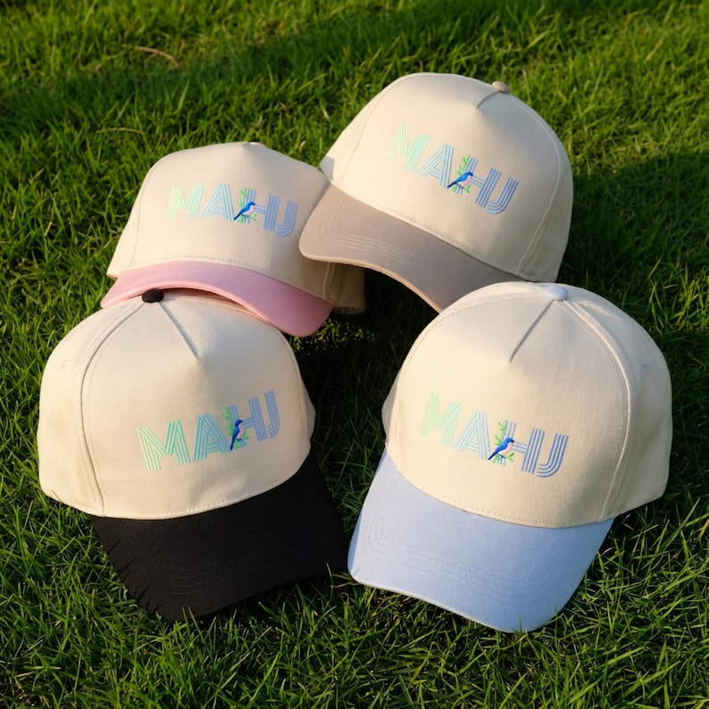 May include: Four baseball caps with cream-colored crowns and brims in pink, beige, black, and light blue. Each cap features the text MAHJ embroidered in a gradient of green to blue, with a small blue bird perched on the letter H.