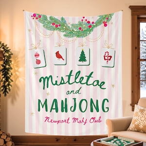 May include: A rectangular wall tapestry featuring a light pink and white vertical striped background. It displays a green pine garland with red berries, four hanging mahjong tiles with holiday icons, and the text Mistletoe and MAHJONG Newport Mahj Club.