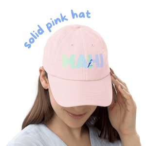 May include: A solid light pink baseball cap featuring the text "MAHJ" in a striped, multi-colored font across the front panel. The letter "H" in the text incorporates a small, blue bird illustration perched on a green branch element.