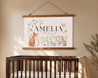 May include: A rectangular wall hanging featuring a white canvas with the name AMELIA Marie printed in black and brown text. The design includes watercolor illustrations of a fox, a rabbit, wildflowers, butterflies, and a snail, suspended by a string.
