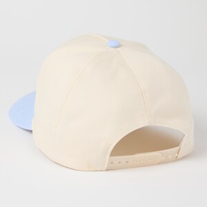 May include: A cream-colored baseball cap featuring a light blue brim and a matching light blue button at the crown. The cap includes an adjustable plastic snapback closure in a cream color, providing a customizable fit for the wearer.