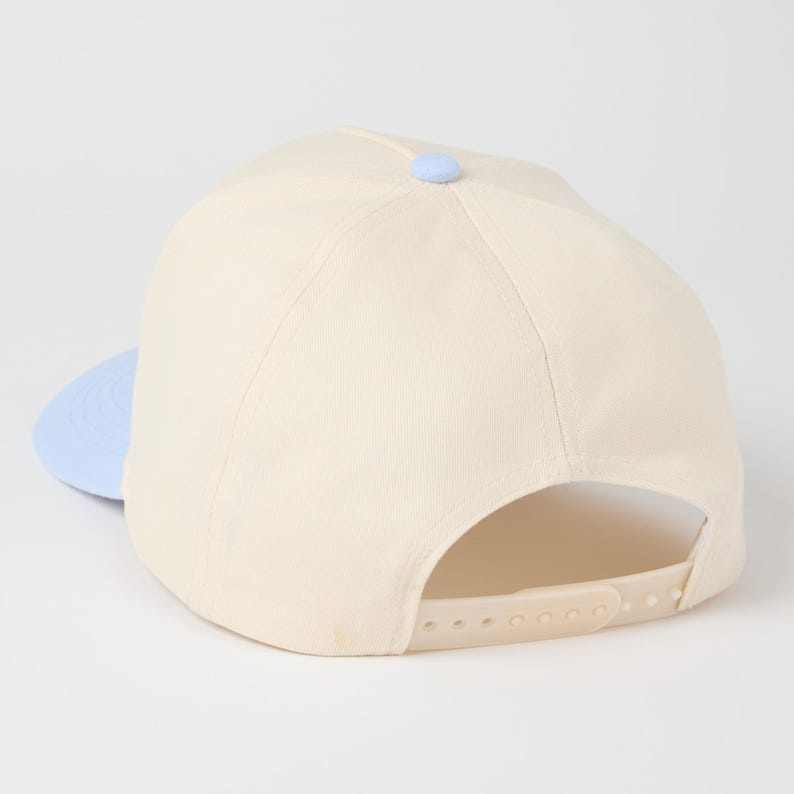 May include: A cream-colored baseball cap featuring a light blue brim and a matching light blue button at the crown. The cap includes an adjustable plastic snapback closure in a cream color, providing a customizable fit for the wearer.