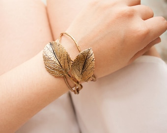 May include: Gold-toned leaf cuff bracelet. The bracelet features two large, detailed leaf designs. The leaves are connected by a thin, curved band that wraps around the wrist. The bracelet is worn on a wrist, showcasing the jewelry's design and color.