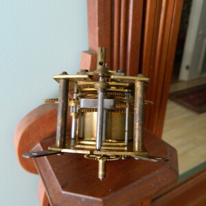 May include: Close-up of a gold-toned clock mechanism with gears and a metal rod. The mechanism is mounted on a wooden surface.