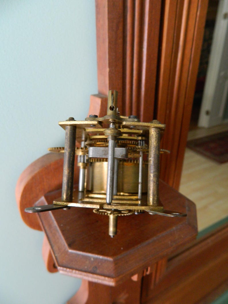 May include: Close-up of a gold-toned clock mechanism with gears and a metal rod. The mechanism is mounted on a wooden surface.