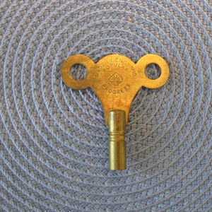 May include: A vintage brass clock key with two circular holes and a cylindrical stem. The key is stamped with the words "MADE IN ENGLAND POPULAR PROGRESS" and a spade symbol. The key is set against a blue woven background.
